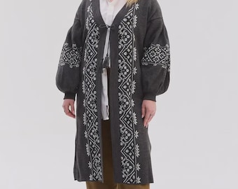 May include: A long, dark gray open-front cardigan featuring white geometric embroidery along the front panels and sleeves. The garment has voluminous sleeves with ribbed cuffs, a tie closure at the neckline with tassels, and a straight hemline.