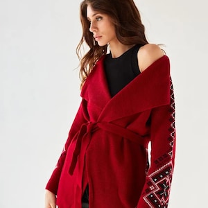 May include: A deep red knit cardigan featuring a wide collar, a matching tie belt at the waist, and long sleeves adorned with intricate black and white geometric patterns running down the outer arms, worn over a simple black sleeveless top.
