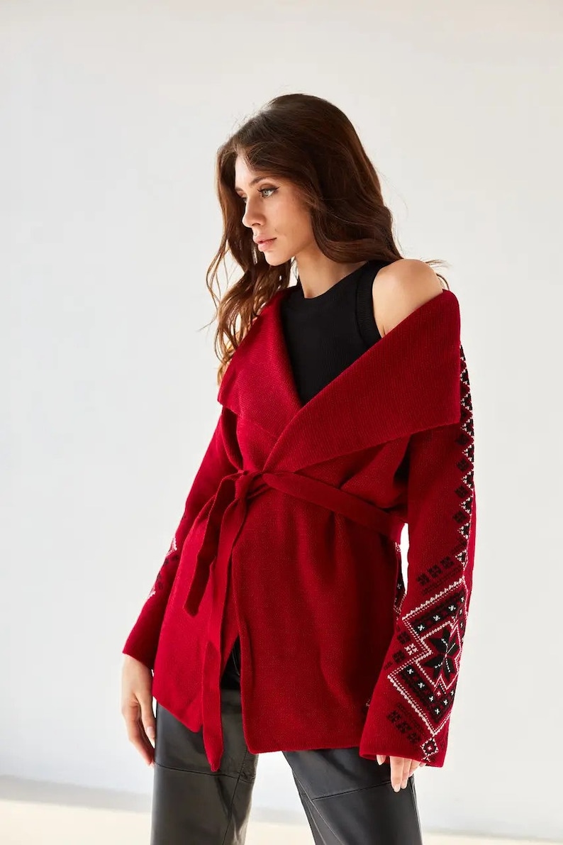 May include: A deep red knit cardigan featuring a wide collar, a matching tie belt at the waist, and long sleeves adorned with intricate black and white geometric patterns running down the outer arms, worn over a simple black sleeveless top.