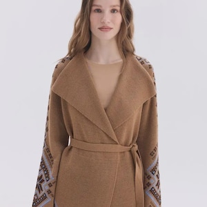 May include: A tan knit cardigan featuring a wide shawl collar and a matching tie belt at the waist. The long sleeves are decorated with a geometric pattern in shades of brown, blue, and white, extending from the shoulders to the cuffs.