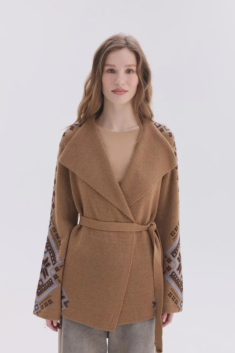 May include: A tan knit cardigan featuring a wide shawl collar and a matching tie belt at the waist. The long sleeves are decorated with a geometric pattern in shades of brown, blue, and white, extending from the shoulders to the cuffs.
