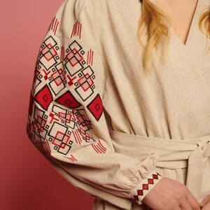 May include: A beige linen dress with red and black embroidered geometric patterns on the sleeves and cuffs. The dress has a V-neckline and a tie belt.