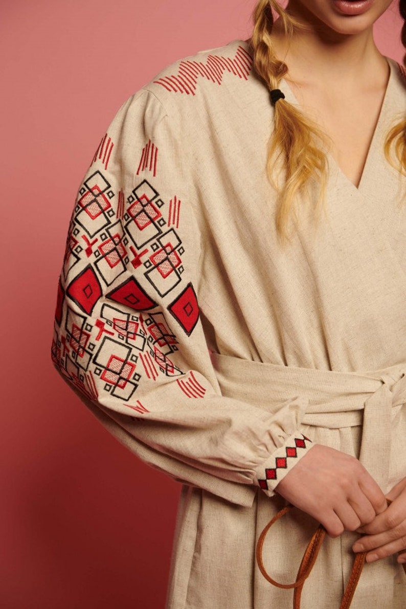 May include: A beige linen dress with red and black embroidered geometric patterns on the sleeves and cuffs. The dress has a V-neckline and a tie belt.