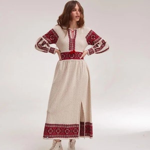 May include: A long, white knitted dress with red embroidered details. The dress has a round neckline, long sleeves, and a fitted waist. The skirt is flowy and has a slit in the front. The model is wearing white sneakers.