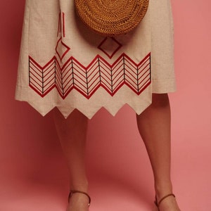 May include: A woman wearing a beige linen skirt with red and black geometric embroidery and a round woven bag. She is wearing beige leather sandals with ankle straps.