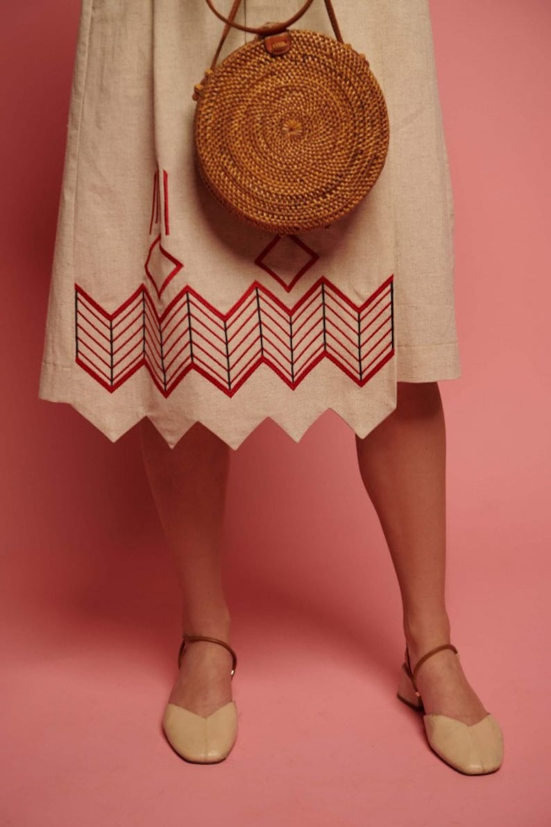 May include: A woman wearing a beige linen skirt with red and black geometric embroidery and a round woven bag. She is wearing beige leather sandals with ankle straps.