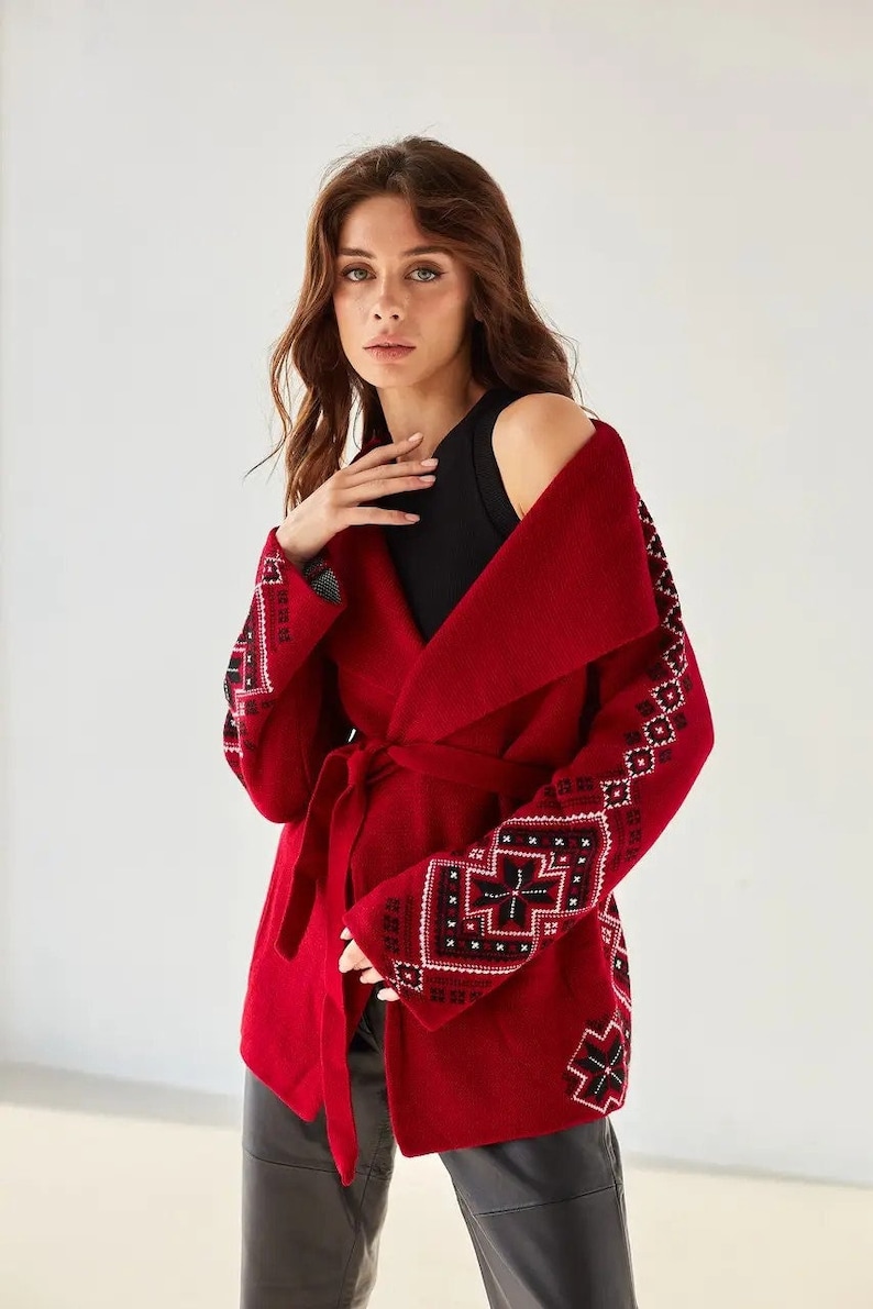 May include: A vibrant red wrap cardigan featuring a wide collar and a matching tie belt. The sleeves and lower sections are decorated with intricate geometric patterns in black and white, creating a bold contrast against the solid red fabric.