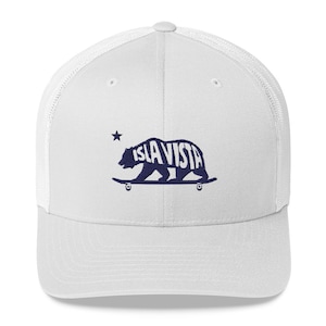 May include: White trucker hat with a navy blue graphic of a bear on a skateboard, with the text "ISLA VISTA" inside the bear's silhouette. A small star is above the bear. The hat has a white mesh back and a curved brim.