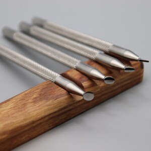 Pu&ograve; includere: Set di cinque strumenti in metallo argentato con punte di forme diverse, disposti in un supporto in legno.