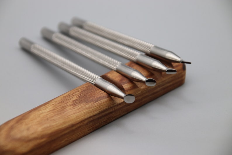 Pu&ograve; includere: Set di cinque strumenti in metallo argentato con punte di forme diverse, disposti in un supporto in legno.