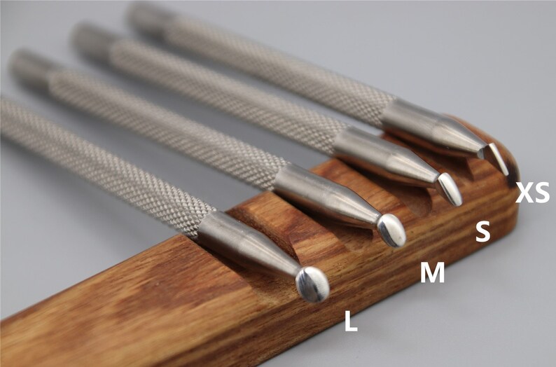 Pu&ograve; includere: Set di quattro strumenti in acciaio inossidabile con punte arrotondate di diverse dimensioni, etichettati XS, S, M ed L, esposti su un supporto in legno.