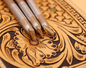May include: Three silver leather tooling stamps with a textured surface, resting on a brown and tan leather background with a floral design.