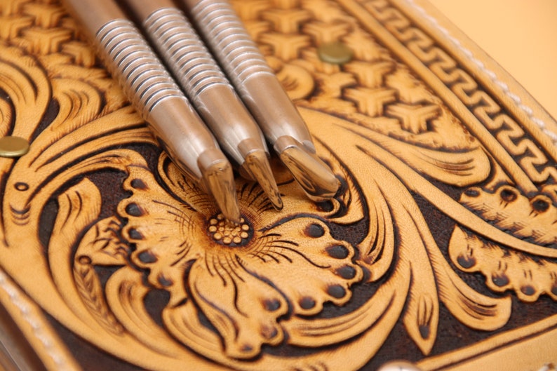 May include: Three silver leather tooling stamps with a textured surface, resting on a brown and tan leather background with a floral design.