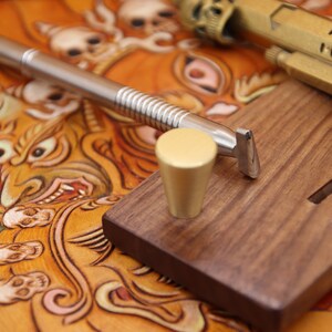 May include: A close-up of a leather carving tool with a wooden handle and a metal blade. The tool is resting on a wooden block with a groove cut into it. A gold-colored metal knob is also visible on the block.