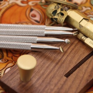 Pu&ograve; includere: Quattro strumenti di lavorazione del cuoio argentati con manici in legno, un accendino in ottone con la scritta "DUSELF" e una manopola in ottone su un blocco di legno.