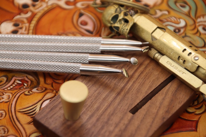 Pu&ograve; includere: Quattro strumenti di lavorazione del cuoio argentati con manici in legno, un accendino in ottone con la scritta "DUSELF" e una manopola in ottone su un blocco di legno.