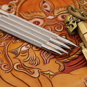 Pu&ograve; includere: Quattro strumenti in argento per la lavorazione del cuoio con manici testurizzati, che giacciono su una superficie in pelle marrone con un design dettagliato. Un portachiavi a forma di teschio dorato e un accendino dorato sono anche sulla superficie.