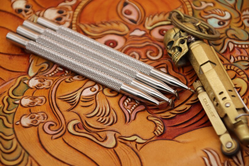 Pu&ograve; includere: Quattro strumenti in argento per la lavorazione del cuoio con manici testurizzati, che giacciono su una superficie in pelle marrone con un design dettagliato. Un portachiavi a forma di teschio dorato e un accendino dorato sono anche sulla superficie.