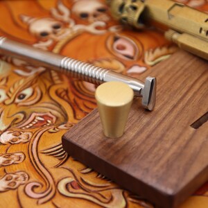 May include: A close-up of a leather working tool set. The set includes a metal tool with a handle and a wooden base. The base has a groove for the tool and a small knob on the side.