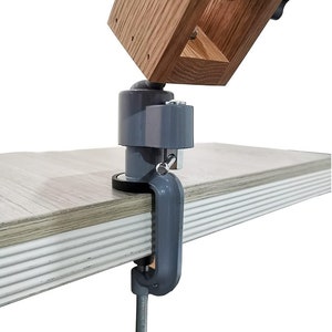 K&ouml;nnte beinhalten: Eine graue Metallklemme mit einem schwarzen Knopf, befestigt an einem Holzarm mit einem grauen Kugelgelenk. Die Klemme ist auf einer hellbraunen Holzoberfl&auml;che montiert.