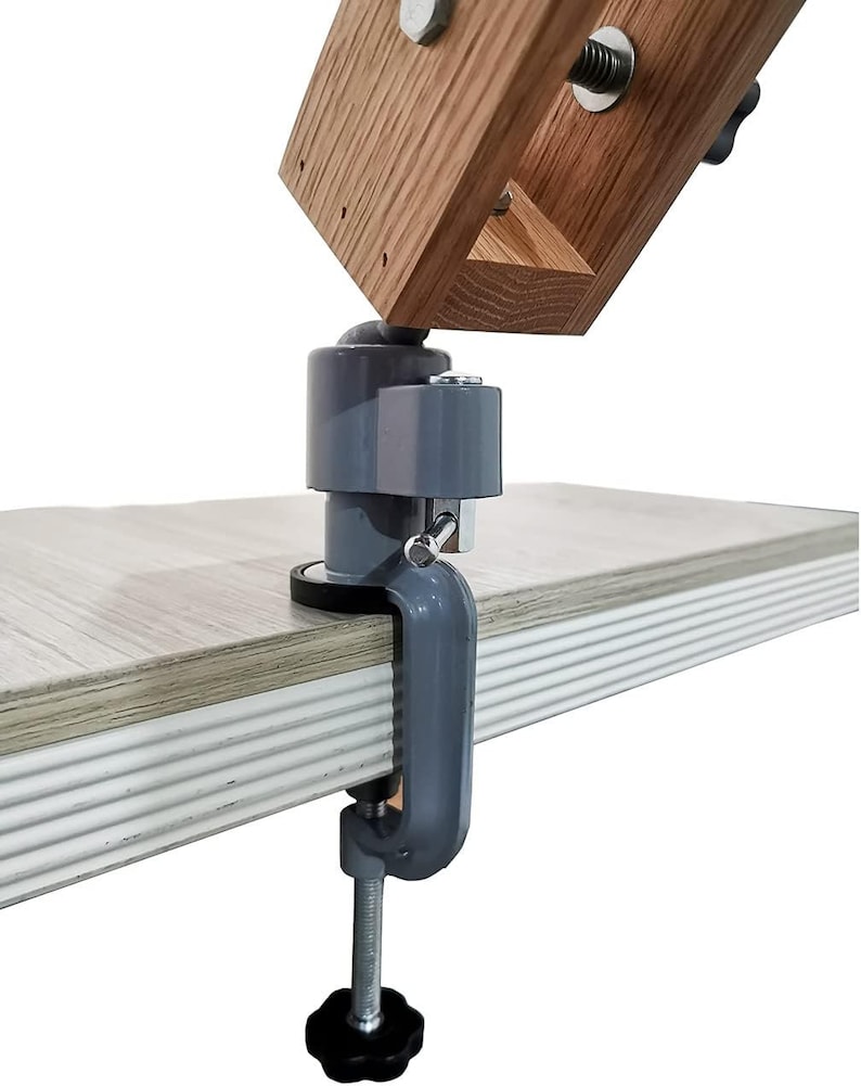 K&ouml;nnte beinhalten: Eine graue Metallklemme mit einem schwarzen Knopf, befestigt an einem Holzarm mit einem grauen Kugelgelenk. Die Klemme ist auf einer hellbraunen Holzoberfl&auml;che montiert.
