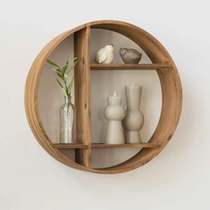 May include: A round, wooden wall shelf with a natural finish. It features three shelves and a vertical divider. The shelf is decorated with two bird figurines, two gray candles, and a clear glass bottle with greenery.