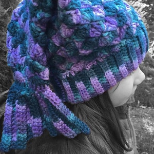 May include: A purple and teal crochet hat with a mermaid tail design. The hat is being worn by a person with long dark hair. The text "MERMAID TAIL HAT CROCHET PATTERN" is displayed below the image.
