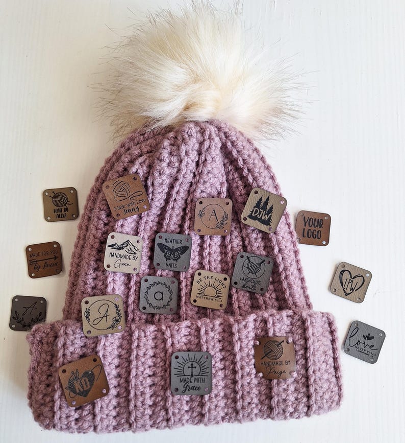 K&ouml;nnte beinhalten: Eine rosafarbene H&auml;kelm&uuml;tze mit einem Kunstfellbommel und verschiedenen Lederpatches mit unterschiedlichen Designs und Texten, darunter "Made with Love Jenny", "Handmade by Grace", "Heather Knits", "DJW", "Your Logo", "Love Never Fails", "Make Path with Grace", "Handmade by Prige" und "A".