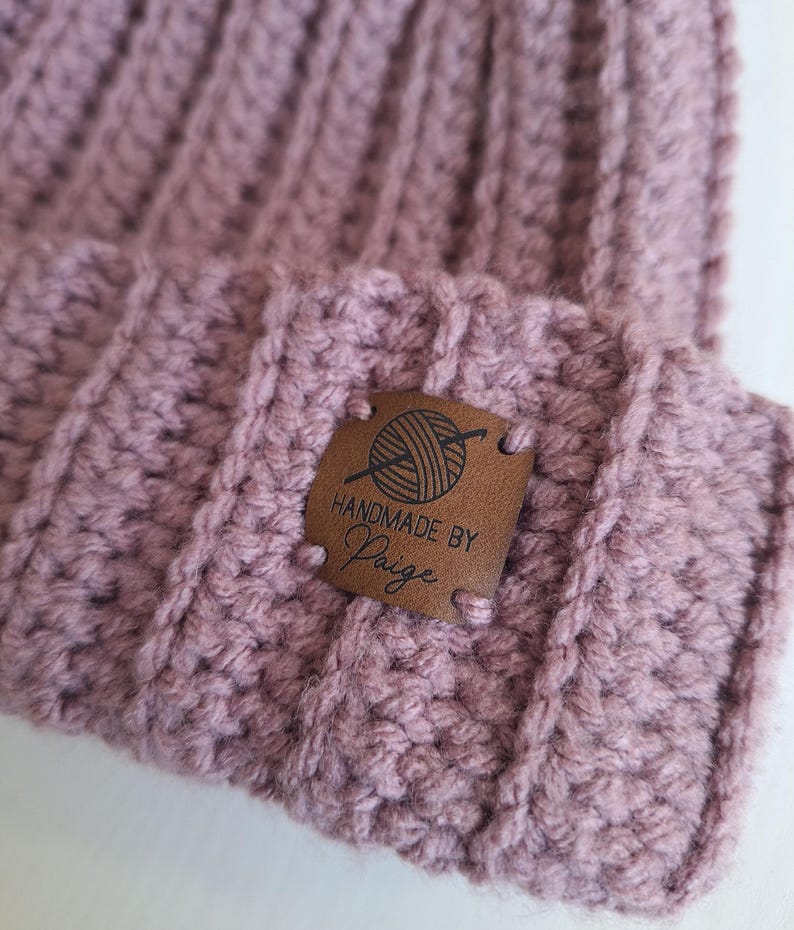 K&ouml;nnte beinhalten: Eine mauvefarbene H&auml;kelm&uuml;tze mit einem braunen Lederanh&auml;nger, auf dem "Handmade by Paige" steht und ein kleines Bild von einem Wollkn&auml;uel und Stricknadeln.
