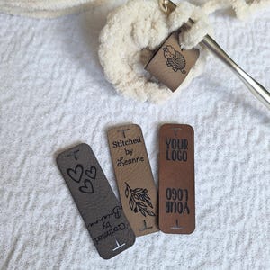 May include: Three rectangular faux leather labels in dark brown, tan, and medium brown, featuring engraved text and icons. One label reads Stitched by Leanne, another Crocheted by Brianna, and the third YOUR LOGO. A crochet hook is inserted into cream yarn.
