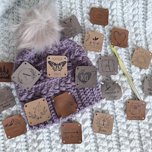 Peut inclure: Un bonnet en tricot violet avec un pompon en fausse fourrure, entouré de diverses étiquettes carrées en cuir. Les étiquettes présentent des motifs gravés, dont un papillon, des initiales et du texte comme "Handmade by Paige" et "Made with Grace". Un crochet est également présent.