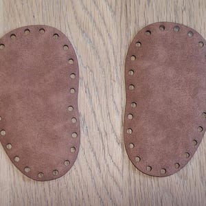 May include: Two brown, oval-shaped leather bases for crochet projects. Each base has a series of evenly spaced holes around the perimeter for attaching yarn. The bases are displayed on a light brown wooden surface.