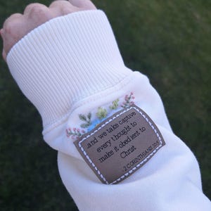 May include: A cream-coloured sweatshirt sleeve featuring a rectangular brown patch with white stitching. The patch is embroidered with the text: and we take captive every thought to make it obedient to Christ - 2 CORINTHIANS 10:5, accented by floral embroidery.
