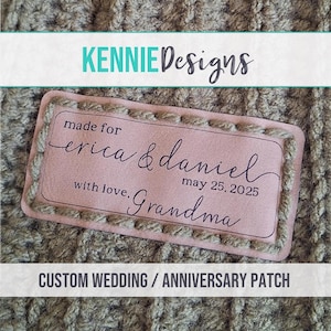 May include: A rectangular light pink leather patch with dark brown text that reads made for erica & daniel may 25, 2025 with love, Grandma. The patch features a stitched border and is attached to a textured brown knitted fabric surface.