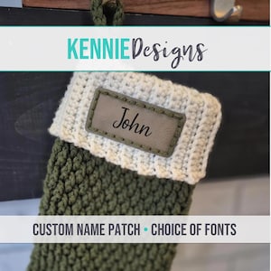 May include: A crocheted olive green Christmas stocking with a white ribbed cuff. A rectangular grey leather-like patch is stitched onto the cuff, featuring the name John in black script. Text reads KENNIE DESIGNS and CUSTOM NAME PATCH.