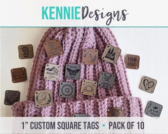 May include: A mauve knit beanie with a white pom-pom, decorated with various 2.54 cm square custom tags featuring engraved designs like mountains, hearts, yarn balls, and text. The tags are scattered across the hat, showcasing personalised branding options.