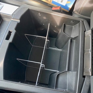 May include: A black plastic car center console organizer with clear plastic dividers. The organizer has multiple compartments for storing items.