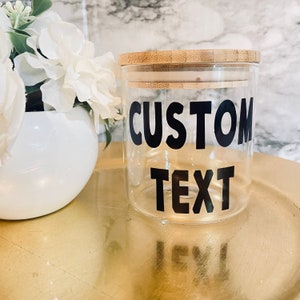 May include: A clear glass jar with a bamboo lid, featuring the words "CUSTOM TEXT" in bold black letters. The jar sits on a gold surface, with a white vase of flowers in the background.