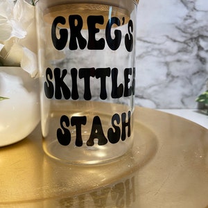 May include: A clear glass jar with a bamboo lid, featuring the text "GREG'S SKITTLES STASH" in black, retro-style lettering. The jar sits on a gold-colored plate, with a white floral arrangement in the background.