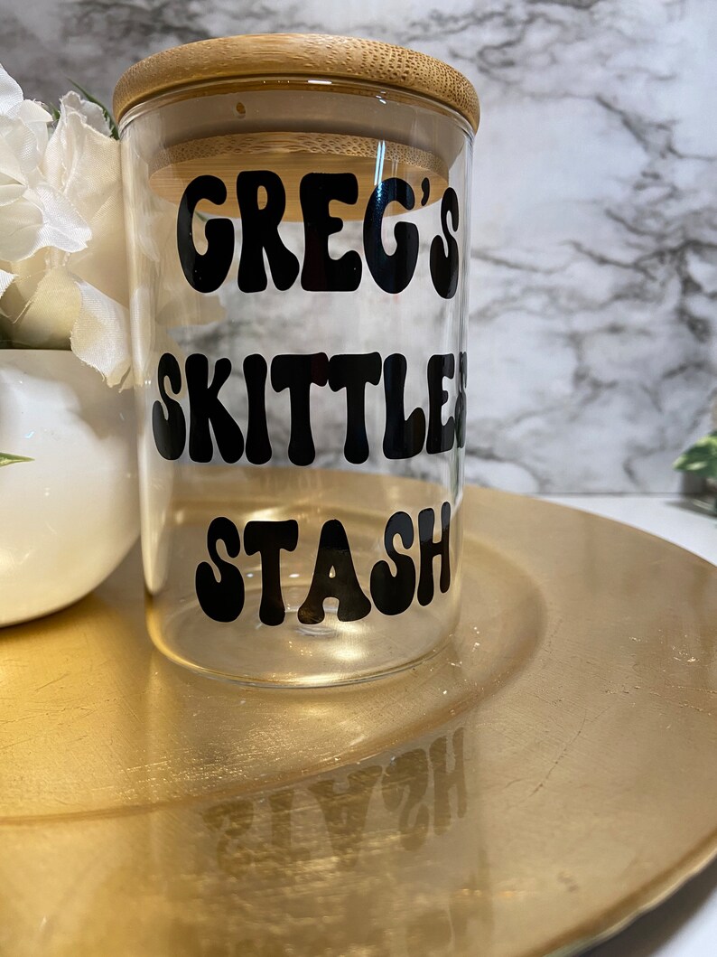 May include: A clear glass jar with a bamboo lid, featuring the text "GREG'S SKITTLES STASH" in black, retro-style lettering. The jar sits on a gold-colored plate, with a white floral arrangement in the background.