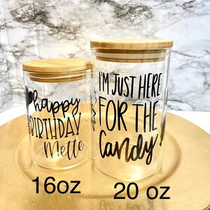 May include: Two glass jars with bamboo lids. The smaller jar is 16 ounces and says "happy birthday Mette". The larger jar is 20 ounces and says "I'm just here for the candy".