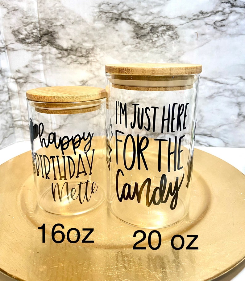 May include: Two glass jars with bamboo lids. The smaller jar is 16 ounces and says "happy birthday Mette". The larger jar is 20 ounces and says "I'm just here for the candy".