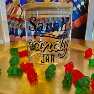 May include: A clear glass jar with a bamboo lid and a black vinyl sticker that says "Sarah's candy JAR". The jar is surrounded by green and red gummy bears on a gold surface.