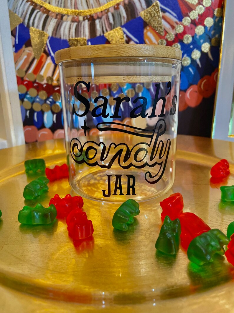 May include: A clear glass jar with a bamboo lid and a black vinyl sticker that says "Sarah's candy JAR". The jar is surrounded by green and red gummy bears on a gold surface.
