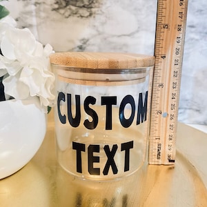 May include: A clear glass jar with a bamboo lid, featuring the words "CUSTOM TEXT" in bold black letters. A wooden ruler is placed next to the jar. The jar is approximately 4 inches tall. White flowers are in the background.