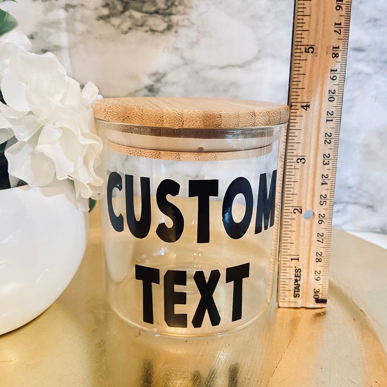 May include: A clear glass jar with a bamboo lid, featuring the words "CUSTOM TEXT" in bold black letters. A wooden ruler is placed next to the jar. The jar is approximately 4 inches tall. White flowers are in the background.