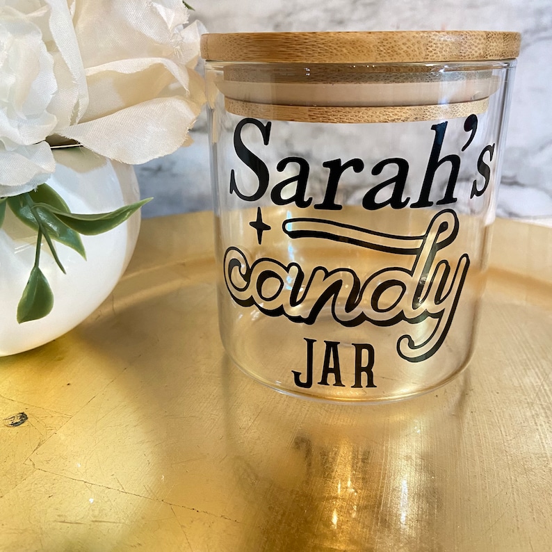 May include: A clear glass jar with a bamboo lid. The jar has a black vinyl decal that says "Sarah's candy JAR" in a retro font.
