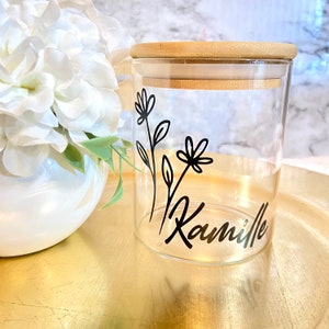 May include: A clear glass jar with a bamboo lid and a black floral design. The jar has the name "Camille" written on it.