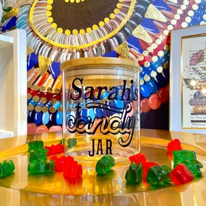 May include: A clear glass jar with a wooden lid and a black decal that says "Sarah's Candy Jar". The jar is sitting on a gold tray with green and red gummy bears.