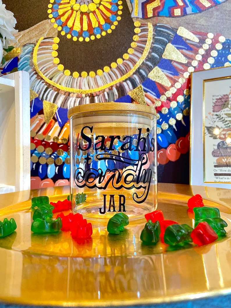 May include: A clear glass jar with a wooden lid and a black decal that says "Sarah's Candy Jar". The jar is sitting on a gold tray with green and red gummy bears.